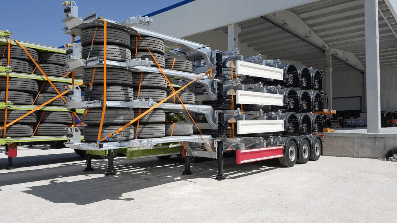 Stacked semi-trailer chassis with tires, secured by orange straps in an outdoor industrial storage area.