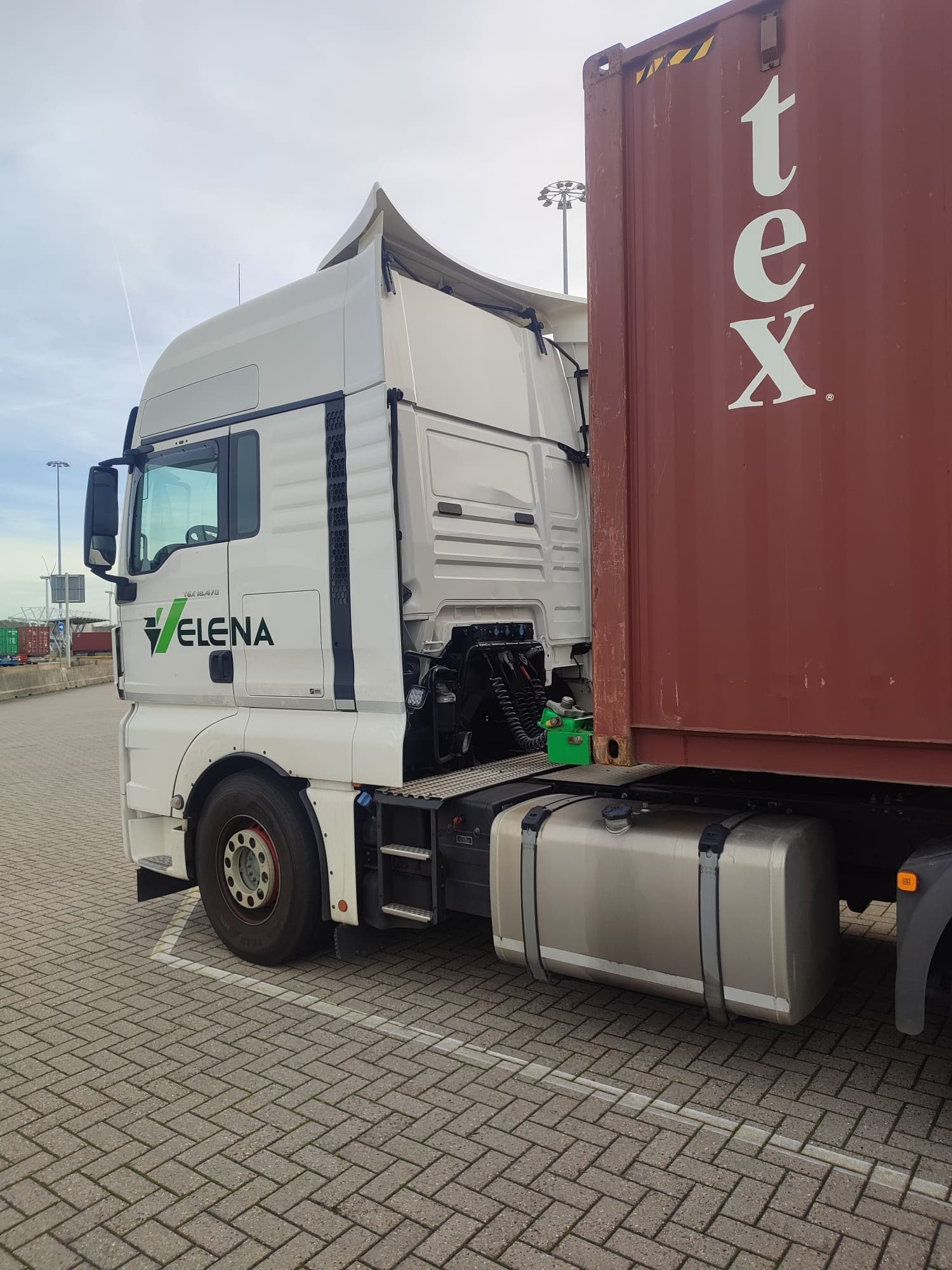 White ELENA semi-truck cab hauling a large red shipping container on a brick-paved lot.